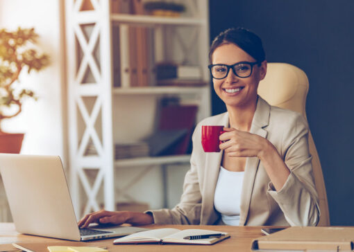 Beautiful expertise. Cheerful young beautiful businesswoman in glasses holding coffee cup and looking at camera with smile while sitting at her working place